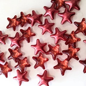 May include: A collection of shiny, star-shaped beads in varying shades of red. The beads have a glossy finish and are scattered across a white surface. The stars have five points and are of similar size.