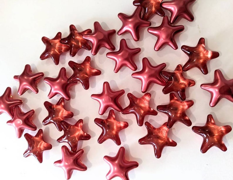 May include: A collection of shiny, star-shaped beads in varying shades of red. The beads have a glossy finish and are scattered across a white surface. The stars have five points and are of similar size.