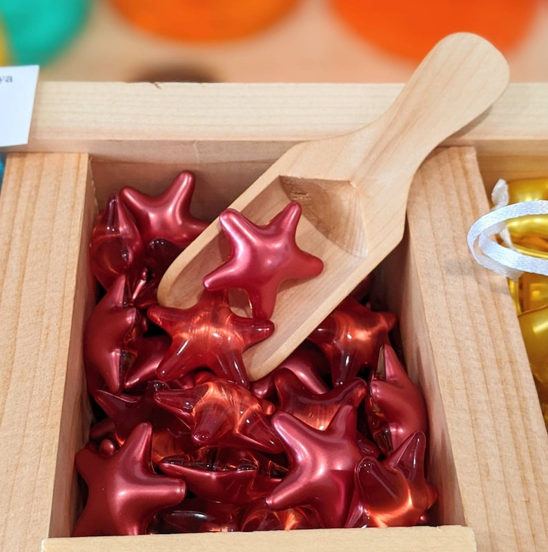 May include: A wooden box containing numerous shiny, red star-shaped items. A small wooden scoop sits amongst the stars. The stars have a glossy finish and appear translucent.