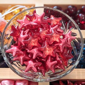 May include: A glass bowl filled with glossy, red star-shaped beads. The beads are clustered together, reflecting light. Other colourful beads are visible in the background, suggesting a craft or decorative theme.