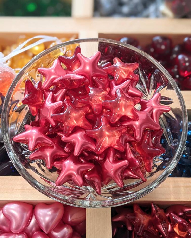 May include: A glass bowl filled with glossy, red star-shaped beads. The beads are clustered together, reflecting light. Other colourful beads are visible in the background, suggesting a craft or decorative theme.
