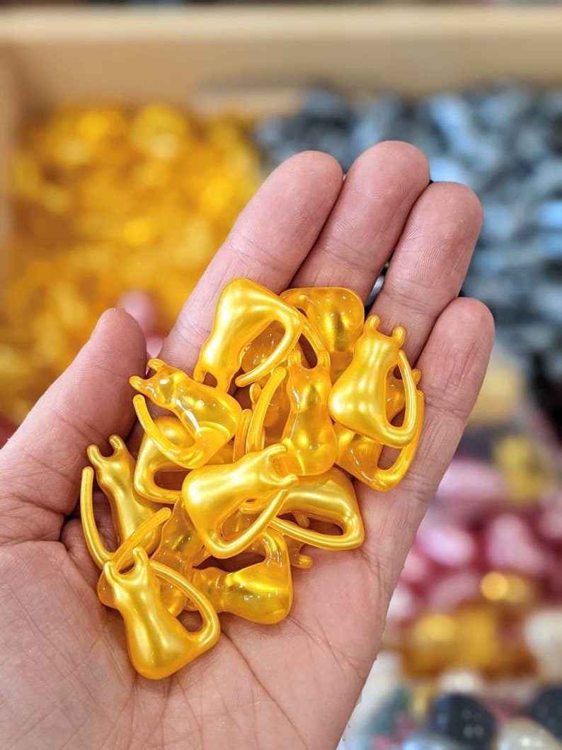 K&ouml;nnte beinhalten: Eine Handvoll kleiner, goldener Kapseln, wahrscheinlich mit einem Sch&ouml;nheitsserum oder einem Nahrungserg&auml;nzungsmittel gef&uuml;llt. Die Kapseln haben die Form kleiner, abgerundeter Figuren.