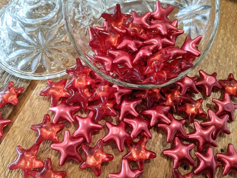 May include: Red star-shaped objects, some in a clear glass bowl and others on a wooden surface. The stars have a glossy finish and a translucent quality. The glass bowl has a decorative, cut-glass design.