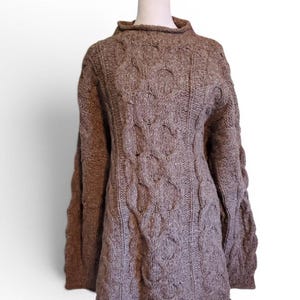 Puede incluir: Un suéter marrón de punto trenzado con cuello alto. El suéter tiene un patrón texturizado y mangas largas. La prenda se exhibe en un maniquí blanco sobre un fondo blanco. El suéter está hecho de un material grueso y cálido.