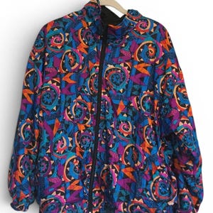 May include: A long-sleeve zip-up jacket featuring a vibrant, multi-colored geometric pattern in shades of blue, purple, orange, and black. The garment includes a high collar, a central black zipper, and elasticized cuffs at the end of each sleeve.