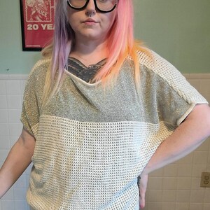 May include: A short-sleeved pullover top featuring a color-blocked design with a metallic gold upper section and a textured, off-white knit lower section. The garment has a wide, relaxed neckline and a ribbed hem, constructed from a lightweight knit fabric.