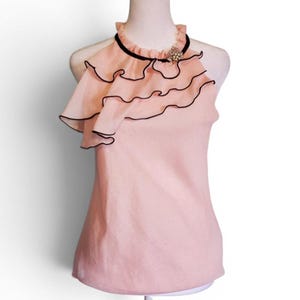 May include: A light pink sleeveless top with a ruffled asymmetrical overlay. The ruffles are outlined in black, and a black band accents the neckline. A decorative brooch is pinned near the neckline. The top is displayed on a mannequin.