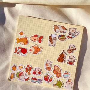 May include: A collection of colorful stickers featuring cartoon fish, cats, and food items like bread, fruit, and sushi. The stickers are arranged on a yellow grid paper, with a soft, off-white background.