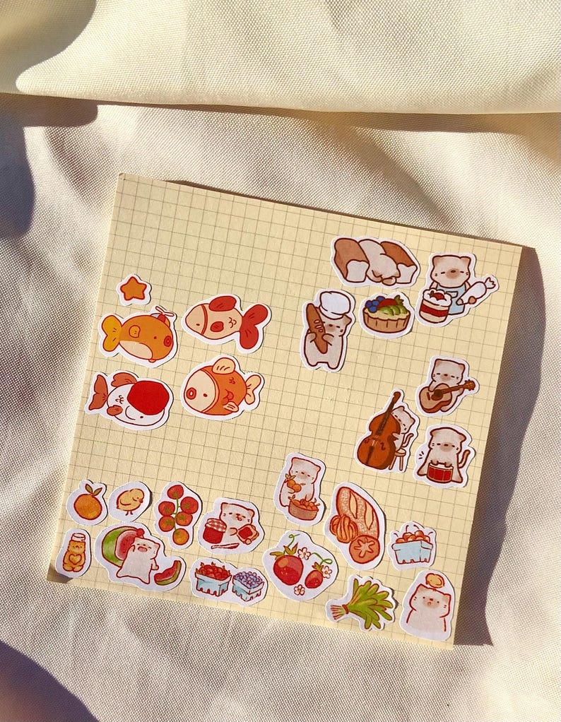 May include: A collection of colorful stickers featuring cartoon fish, cats, and food items like bread, fruit, and sushi. The stickers are arranged on a yellow grid paper, with a soft, off-white background.