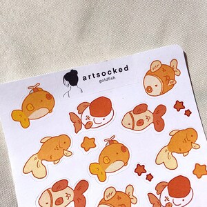 May include: A sheet of stickers featuring cartoon goldfish in shades of orange and white, with star accents. The stickers have a whimsical design, with the text "artsocked goldfish" printed at the top.