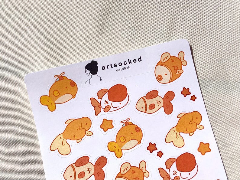 May include: A sheet of stickers featuring cartoon goldfish in shades of orange and white, with star accents. The stickers have a whimsical design, with the text "artsocked goldfish" printed at the top.