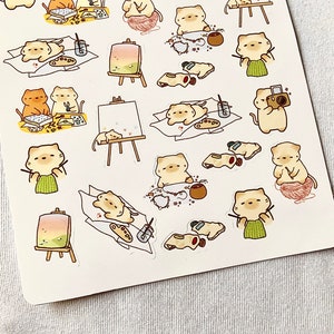 May include: Sheet of stickers featuring cartoon cats engaged in various artistic activities. The cats are depicted painting at easels, knitting with yarn, and taking photos. The stickers have a soft color palette with a white background.