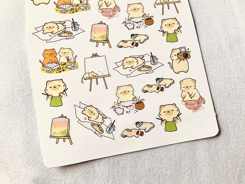 May include: Sheet of stickers featuring cartoon cats engaged in various artistic activities. The cats are depicted painting at easels, knitting with yarn, and taking photos. The stickers have a soft color palette with a white background.