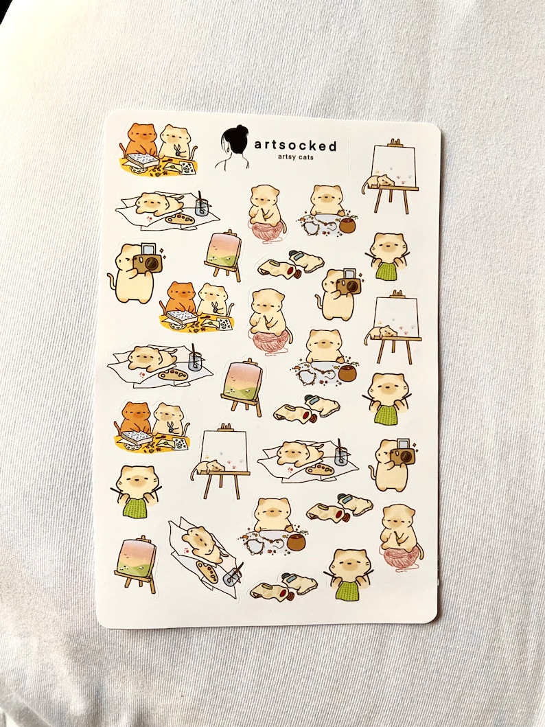 May include: A sticker sheet featuring various illustrations of cats engaged in artistic activities. The cats are depicted painting, taking photos, and relaxing. The sheet includes the text "artsocked" and "artsy cats".