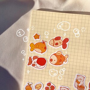 May include: A collection of colorful stickers featuring cartoon fish, a star, and various food items like fruit and jam. The stickers are arranged on a grid-lined paper, with whimsical bubble and star doodles around the edges.