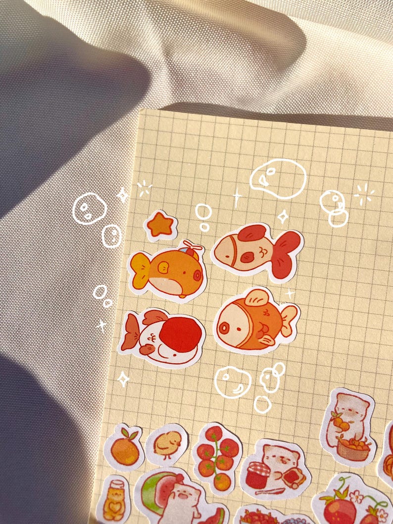 May include: A collection of colorful stickers featuring cartoon fish, a star, and various food items like fruit and jam. The stickers are arranged on a grid-lined paper, with whimsical bubble and star doodles around the edges.