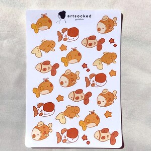 May include: A sheet of stickers featuring cartoon goldfish in various poses and colors, including orange, white, and brown. The stickers have a cute, whimsical design, with some star-shaped accents. The text "artsocked goldfish" is visible.