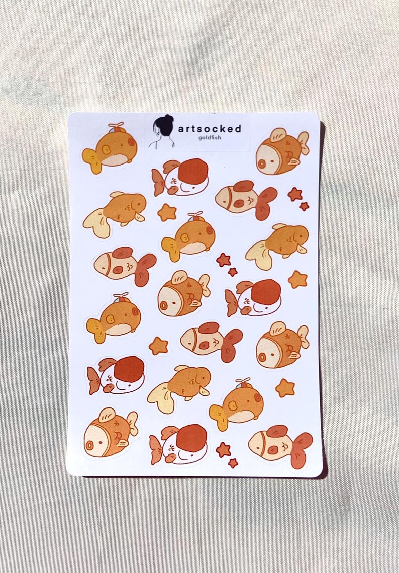 May include: A sheet of stickers featuring cartoon goldfish in various poses and colors, including orange, white, and brown. The stickers have a cute, whimsical design, with some star-shaped accents. The text "artsocked goldfish" is visible.