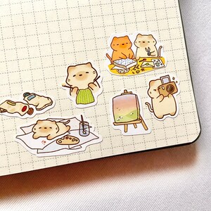 May include: A collection of cute cat stickers in various artistic poses. One cat paints, another photographs, and others create art. The stickers are on a grid paper background, with paint tubes, palettes, and easels.