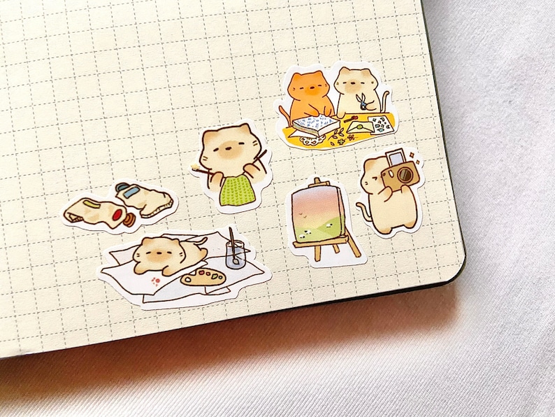 May include: A collection of cute cat stickers in various artistic poses. One cat paints, another photographs, and others create art. The stickers are on a grid paper background, with paint tubes, palettes, and easels.