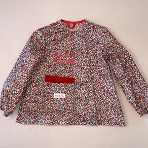 Peut inclure: Une blouse à manches longues avec un motif floral dense rouge, bleu et vert sur fond blanc. Elle présente un col rouge, une poche avant bordée de rouge avec une étiquette blanche portant l'inscription Didine & Anton et un petit drapeau français.