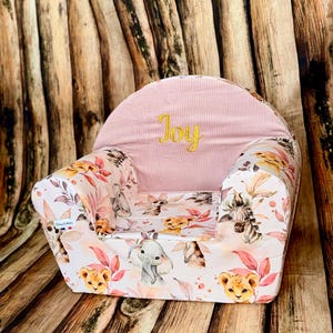 May include: A soft foam armchair featuring a light pink corduroy backrest with the embroidered name Joy in gold thread. The seat and armrests are covered in a white fabric patterned with watercolour illustrations of baby safari animals and leaves.