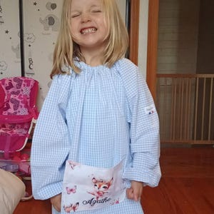 Könnte beinhalten: Hellblau-weiß kariertes Kleid mit langen Ärmeln. Das Kleid hat eine Tasche mit einem Hirsch-Motiv und dem Namen "Agathe". Das Kind lächelt.