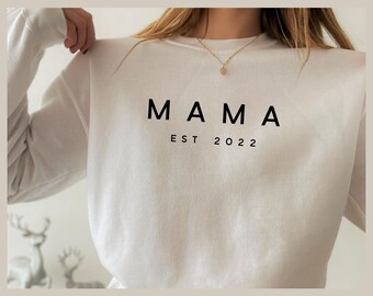 以下が含まれることがあります： 前面に「MAMA EST 2022」とプリントされた白い長袖スウェットシャツです。