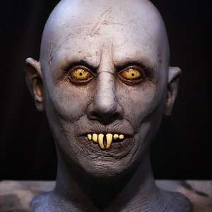 Puede incluir: Primer plano de una cara esculpida detallada de color azul pálido con ojos amarillos y dientes afilados. La cara es parte de una escultura más grande, con el cuello y los hombros visibles.