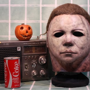 Puede incluir: Una máscara de Halloween blanca con un aspecto desgastado y deteriorado. La máscara tiene agujeros negros para los ojos y una peluca marrón. La máscara está colocada sobre un soporte negro delante de una radio vintage y una lata de Coca-Cola.