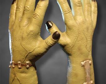 Può includere: Un paio di mani di mostro verdi con unghie marrone scuro. Le mani hanno vene dettagliate e sono adornate da un braccialetto marrone e una placca di metallo. Le mani sono poste su uno sfondo grigio scuro.
