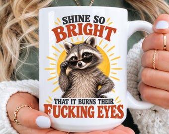 May include: A white ceramic mug featuring a graphic of a raccoon wearing sunglasses in front of a yellow sun. The text reads SHINE SO BRIGHT THAT IT BURNS THEIR FUCKING EYES in bold orange and black lettering, centered on the front of the mug.