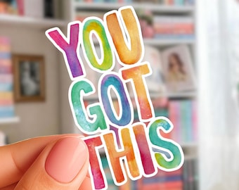 May include: A colorful sticker with the text "YOU GOT THIS" in rainbow hues. The sticker is held in front of a blurred background of bookshelves filled with books.