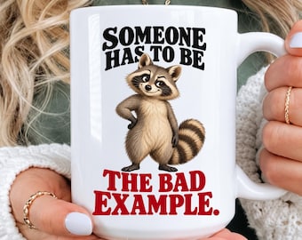 May include: A white ceramic mug featuring a cartoon raccoon illustration centered on the front. Above the raccoon, the black text reads SOMEONE HAS TO BE. Below the raccoon, the red text reads THE BAD EXAMPLE. The mug has a handle on the right side.
