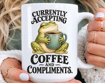 May include: A white ceramic mug featuring a graphic of a green frog holding a small mug with steam rising. The text reads CURRENTLY ACCEPTING COFFEE AND COMPLIMENTS. The mug has a curved handle and is held by hands with gold rings.