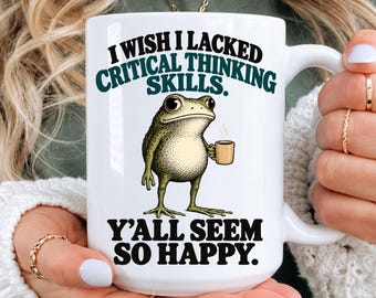 May include: A white ceramic mug featuring a green frog illustration holding a small brown coffee cup. The text above the frog reads I WISH I LACKED CRITICAL THINKING SKILLS. The text below the frog reads Y'ALL SEEM SO HAPPY. The mug has a handle.