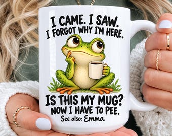 May include: A white ceramic mug featuring a cartoon green frog holding a small white cup. The text reads: I CAME. I SAW. I FORGOT WHY I'M HERE. IS THIS MY MUG? NOW I HAVE TO PEE. See also: Emma. The mug has a curved handle and a smooth, glossy finish.