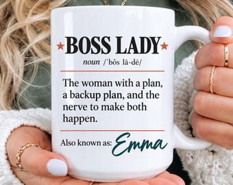 May include: A white ceramic mug featuring the black text BOSS LADY, a phonetic pronunciation, and a definition describing a woman with a plan, a backup plan, and the nerve to make both happen. The text also includes the name Emma in dark green.