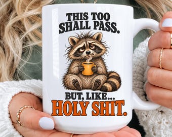 May include: A white ceramic mug featuring a graphic of a tired-looking raccoon holding a small orange coffee cup. The text above reads THIS TOO SHALL PASS and the text below reads BUT, LIKE... HOLY SHIT. The mug has a handle on the right side.