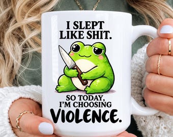 May include: A white ceramic mug featuring a green frog illustration holding a small knife. The text reads: I SLEPT LIKE SHIT. SO TODAY, I'M CHOOSING VIOLENCE. The design is centered on the front of the mug, which has a standard handle and cylindrical shape.