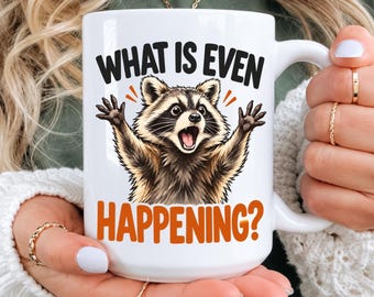 May include: A white ceramic mug featuring a graphic illustration of a surprised raccoon with its paws raised. The text above the raccoon reads WHAT IS EVEN in black, and the text below reads HAPPENING? in orange, set against a white background.