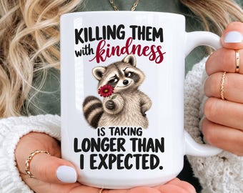 May include: A white ceramic mug featuring a graphic of a raccoon holding a red flower. The text reads: KILLING THEM with kindness IS TAKING LONGER THAN I EXPECTED. The mug has a large handle and is held by two hands with rings on the fingers.