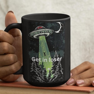 Könnte beinhalten: Schwarze Keramik-Tasse mit einem UFO-Design, einem grünen Lichtstrahl, einem Wald und dem Text "Get in loser". Die Tasse hat einen schwarzen Henkel. Das Bild zeigt eine Tasse, die von einer Person gehalten wird.