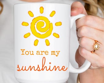 May include: White ceramic mug featuring a yellow sun design with orange accents and the text "You are my sunshine" in orange script. The mug has a large handle and is held by a person.