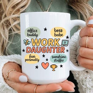 以下が含まれることがあります： カラフルなデザインで「WORK DAUGHTER」の文字が入った白いセラミックマグ。マグには、蜂、ハート、星など、追加のテキストとグラフィックが施されています。マグは、グリーンのシャツと白いセーターを着た人が持っています。