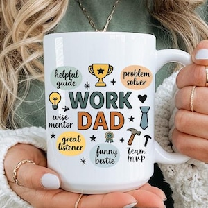 Può includere: Tazza in ceramica bianca con le parole "WORK DAD" e vari attributi positivi come "helpful guide" e "problem solver" in un design colorato. La tazza è tenuta da una persona che indossa una camicia verde e un maglione bianco.