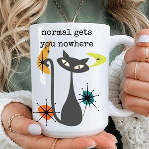 Op de afbeelding: Witte keramische mok met een zwarte kat en een kleurrijk mid-century modern design. De mok heeft de tekst "normal gets you nowhere".