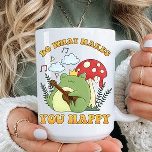 Op de afbeelding: Witte keramische mok met een grillig ontwerp met een kikker die ukulele speelt, een paddenstoel en de tekst "DO WHAT MAKES YOU HAPPY" in een retro lettertype. De mok heeft een groot handvat.