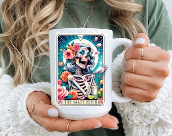 May include: White ceramic mug with a colorful illustration of a skeleton with flowers in its hair. The mug has a black handle and the text "The Salty Bitch" on the front.