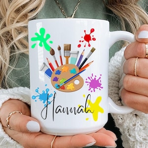Può includere: Tazza in ceramica bianca con un design colorato di tavolozza e pennelli. La tazza ha il nome "Hannah" scritto su di essa.
